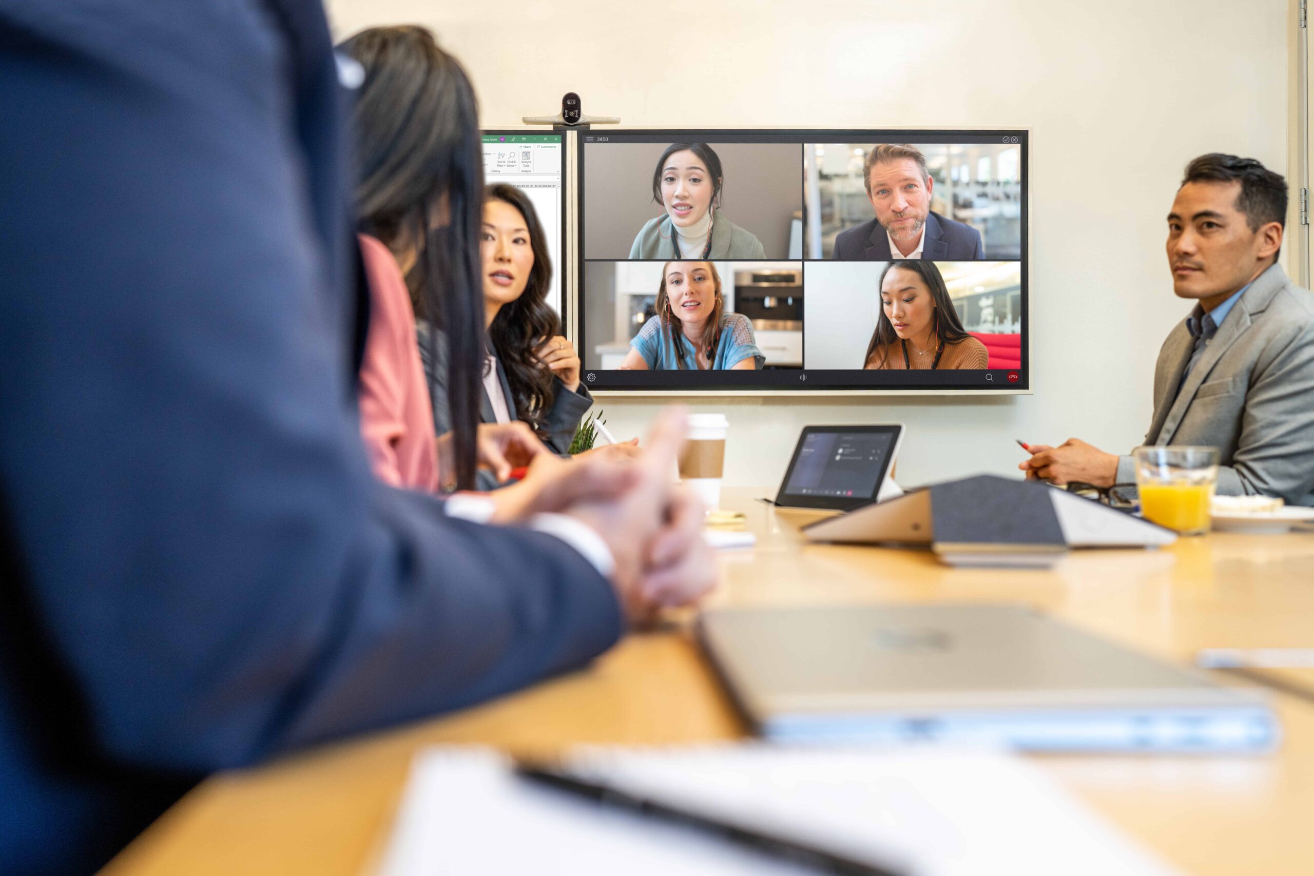 Six people sit at a conference table with laptops, engaging in a video call with four participants visible on the wall screen.