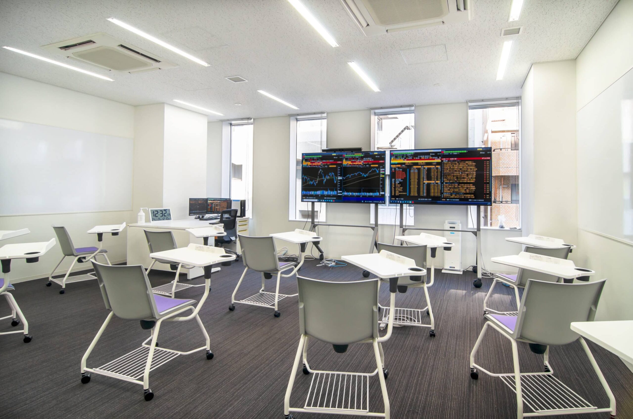 Modern classroom: white desks, gray chairs in rows; screens show graphs. Natural light through tall windows, whiteboard on left wall.