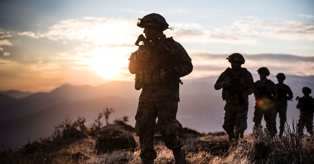 Soldiers with rifles walk a grassy hill at sunrise, mountains in view; dramatic light filters through cloudy sky.