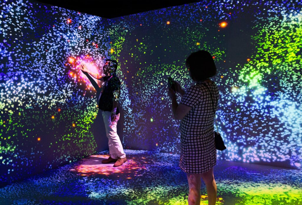 Two people interact with vibrant digital art; one touches the wall as the other takes a photo amid blue, green, and purple hues.