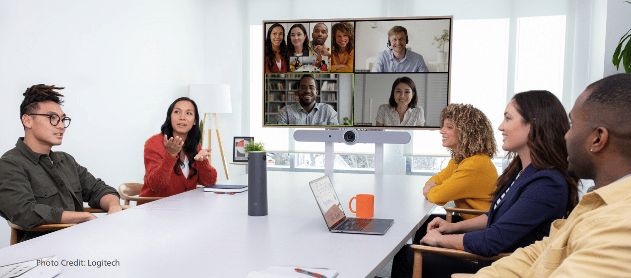 Six people sit around a table with a video call on screen showing seven colleagues. A laptop and speakerphone are on the table.