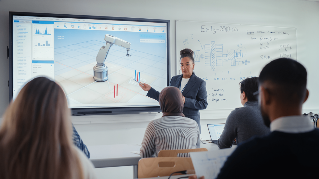 Teacher points at robotic arm simulation; students take notes. Whiteboard with diagrams in background.