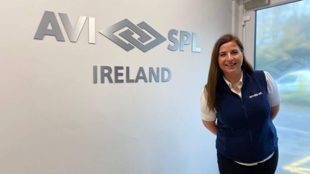 A woman in a white shirt and dark vest smiles by an office lobby wall with the AVI SPL IRELAND logo.