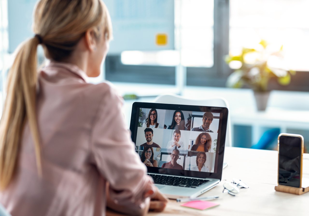 Woman with ponytail video conferencing, nine participants shown; smartphone and office items on desk in bright room.