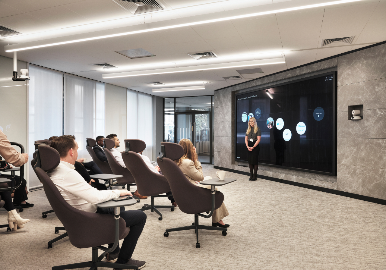 A woman presents data on a large screen in a modern conference room with attendees seated in swivel chairs.