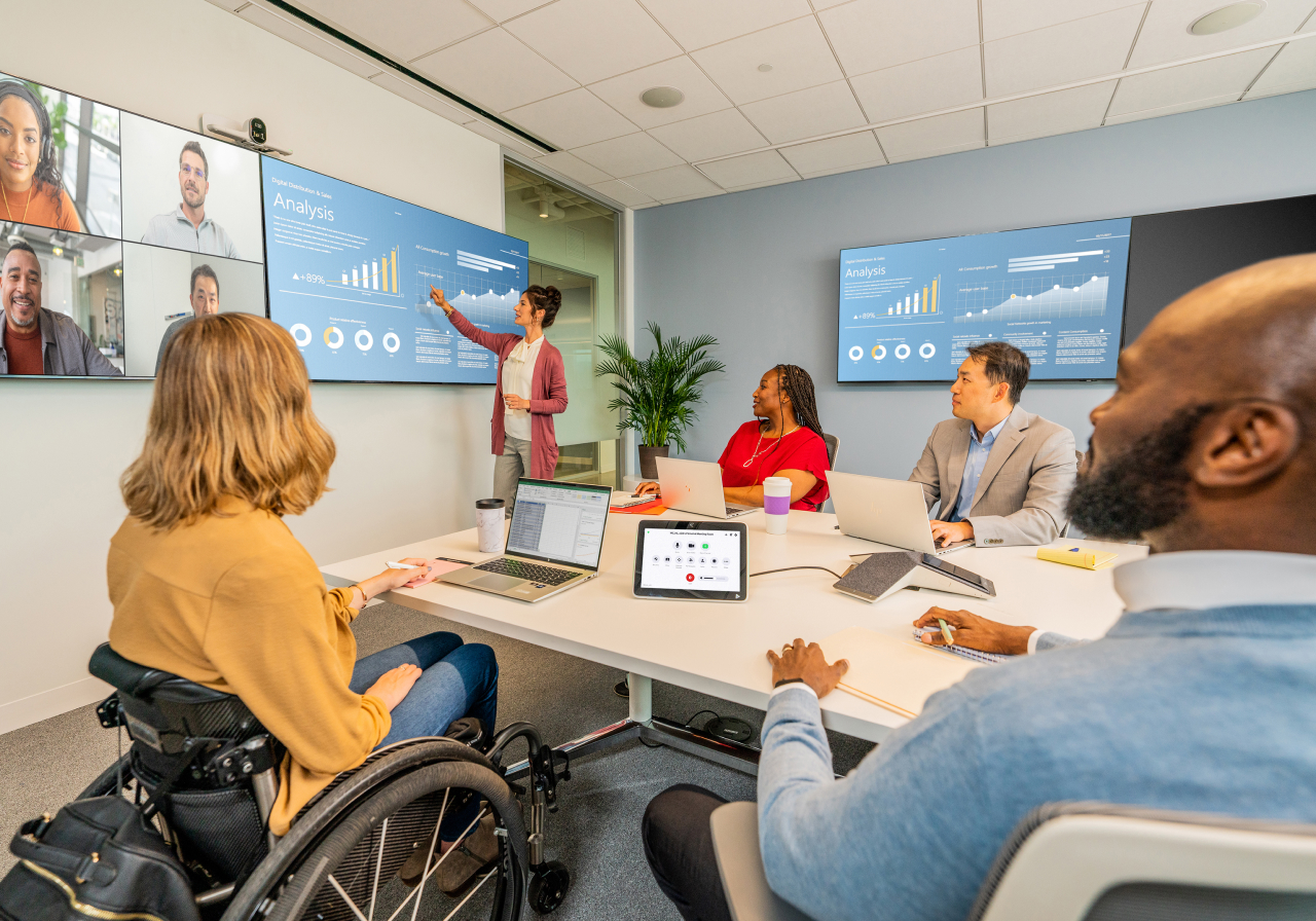 Team in modern room reviews screen charts; presenter, laptops, wheelchair user, virtual attendees on side screen.