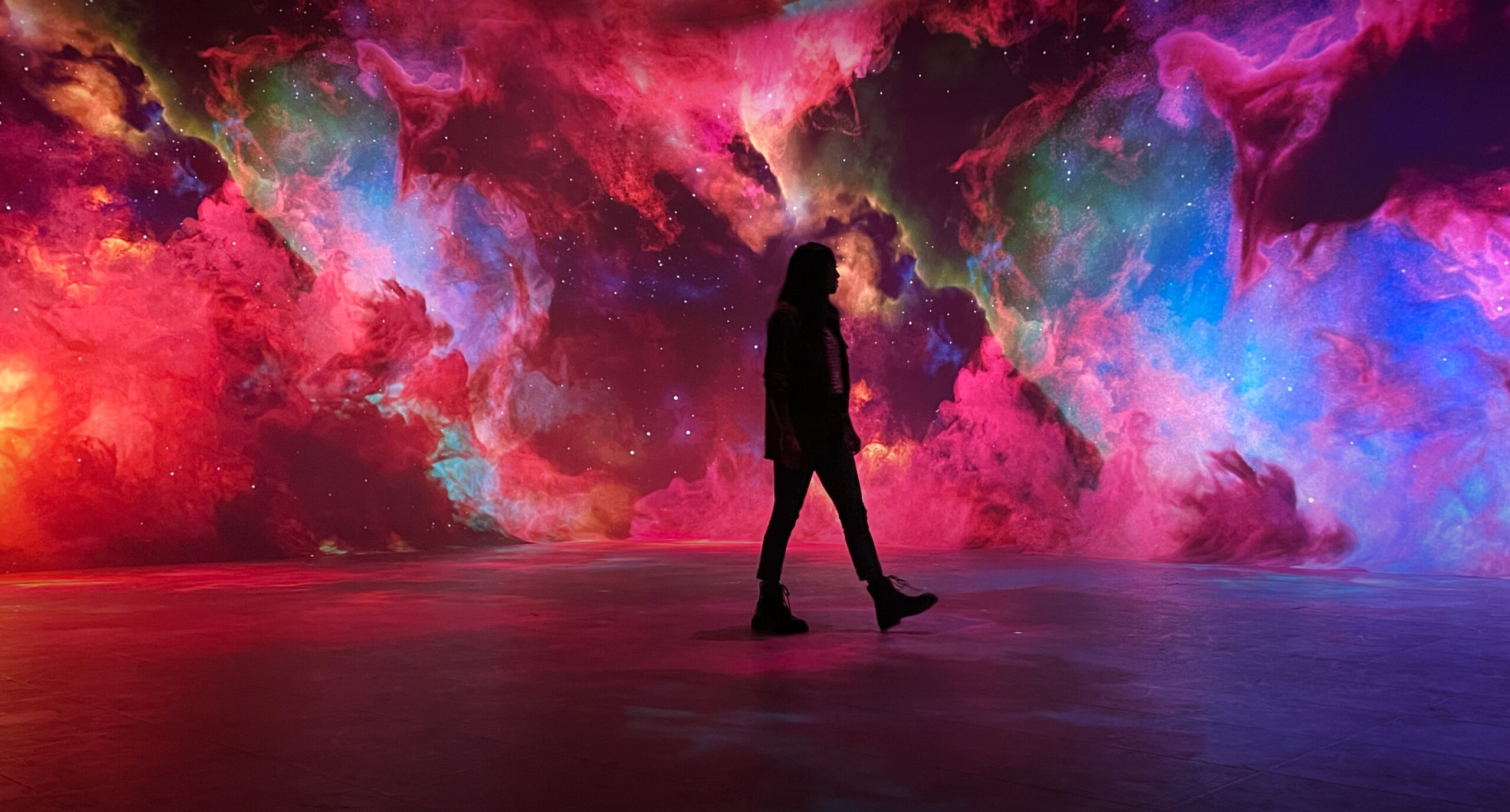 A silhouette walks before a vibrant cosmic nebula of swirling reds, blues, and greens, creating an immersive and surreal scene.