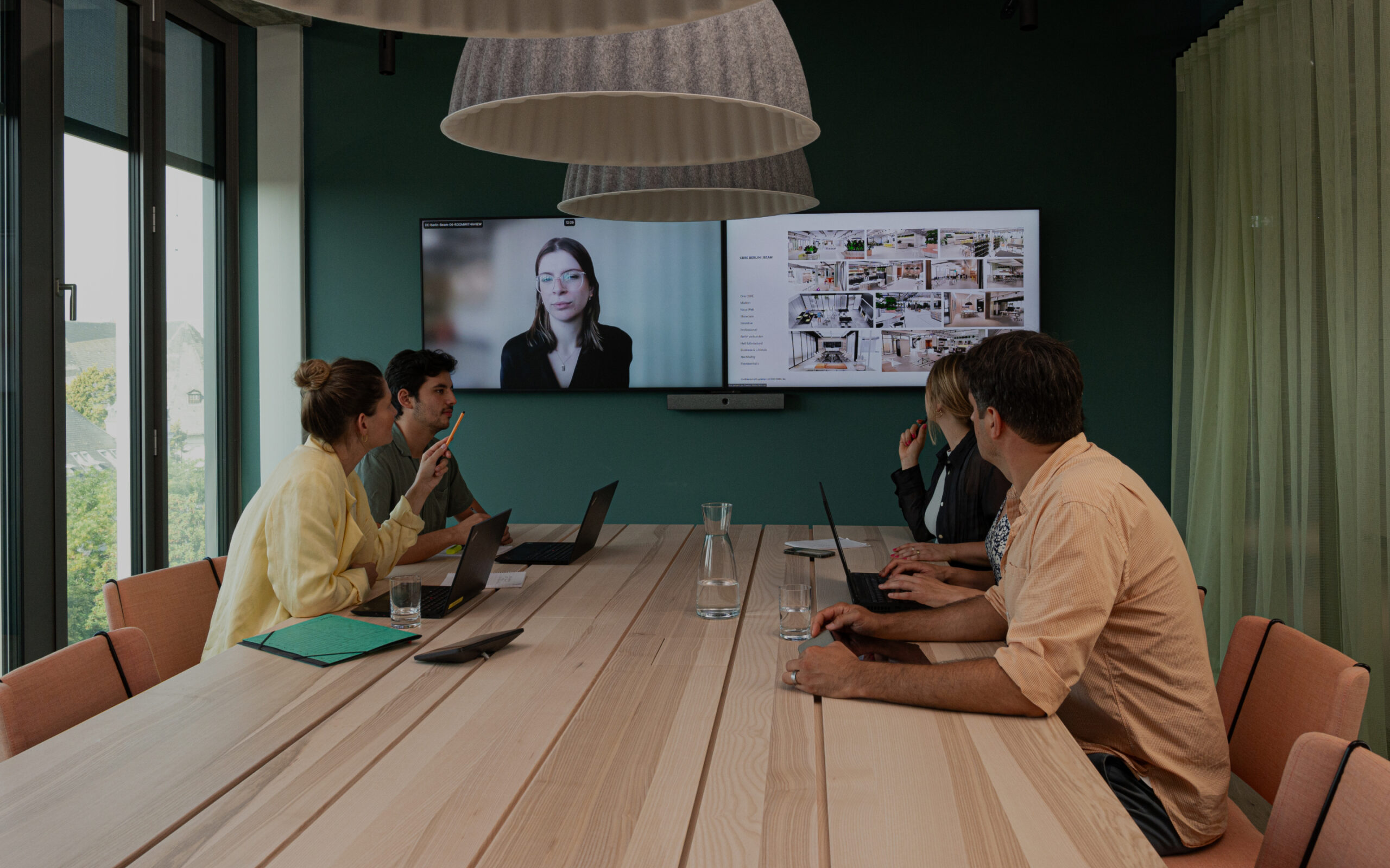 Five people in a meeting room, four seated at a long table with laptops, watching a video call and presentation, natural light floods in.