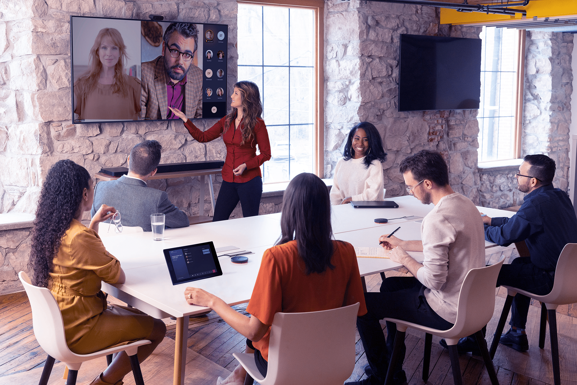 Seven people sit at a table; a woman stands by a screen showing two on a video call. Stone walls, large windows, devices on the table.