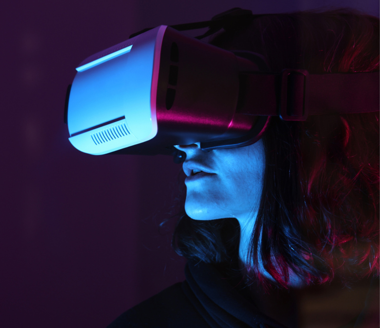 Person wearing a virtual reality headset, illuminated by blue and red lighting, looking upwards. The background is dark, emphasizing the glow and contours of the headset.