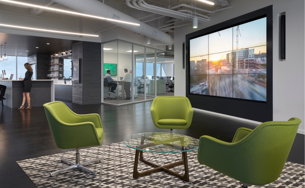 Modern office lounge: green chairs, glass table on patterned rug, large screen cityscape. Woman at counter; two people in glass room.