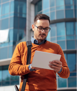 A man in glasses and an orange sweater stands in front of a modern glass building, looking at a tablet. He carries a shoulder bag, and the sun casts a soft light on the scene.