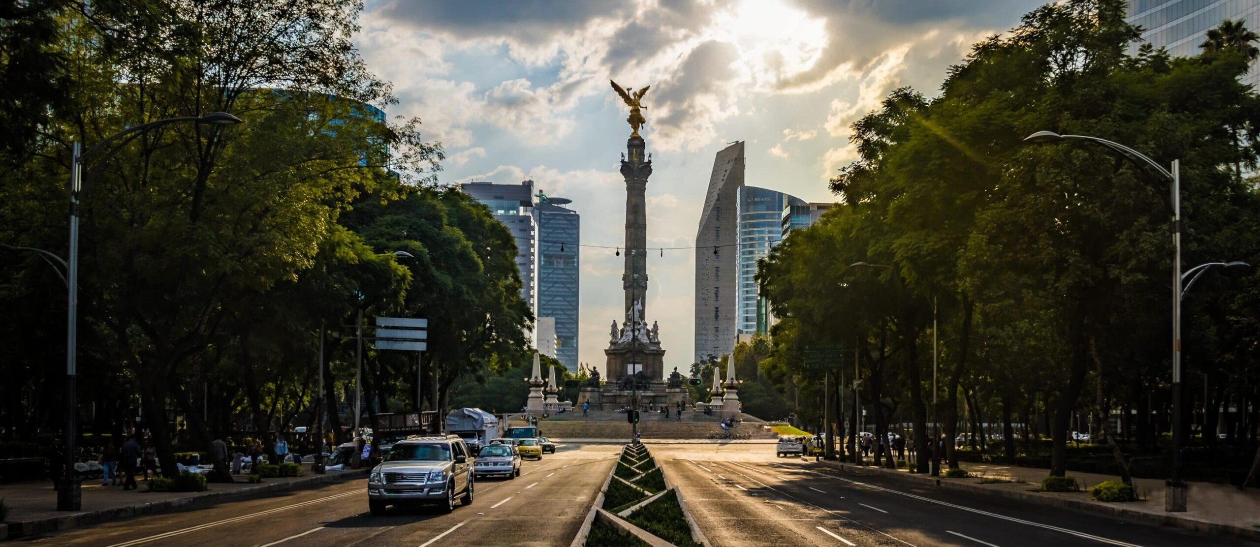 Wide boulevard with a golden-angel monument, tall modern buildings, lush trees, dramatic sky, sunlight beams, cars on road.