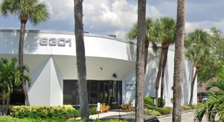 White building 8801 with palm trees, greenery, road in front, and partly cloudy sky.