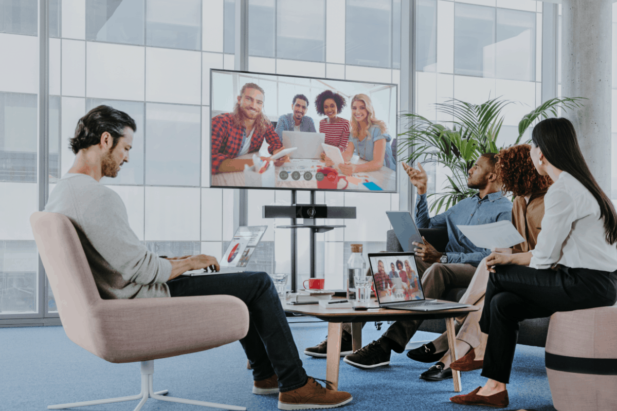 A team of 4 in an office, video conferencing with 4 others. Laptops and documents out, large windows, and a potted plant nearby.