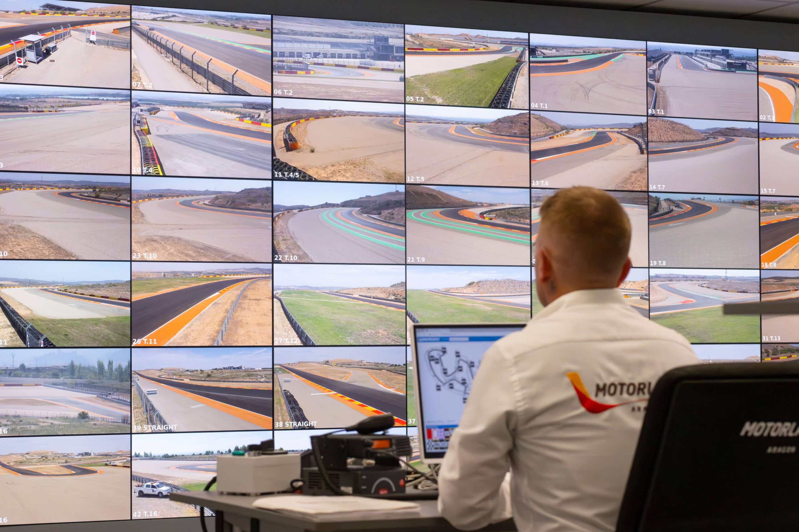 A person in a white shirt monitors racetrack feeds on multiple screens, showing various circuit angles under clear skies.