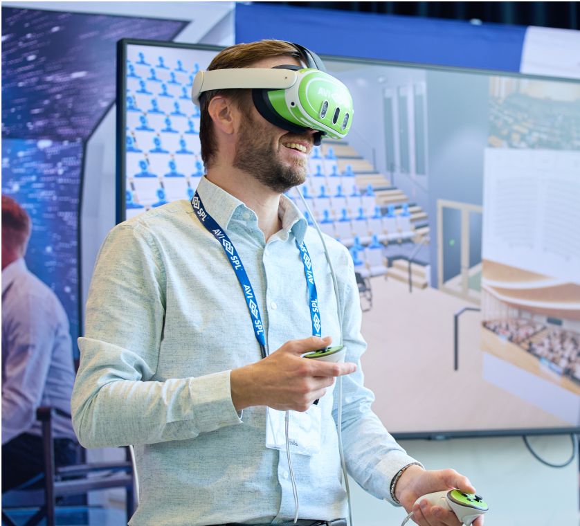 A man with a VR headset smiles, holding controllers. He stands before a virtual auditorium screen, wearing an AVEVA lanyard and badge.