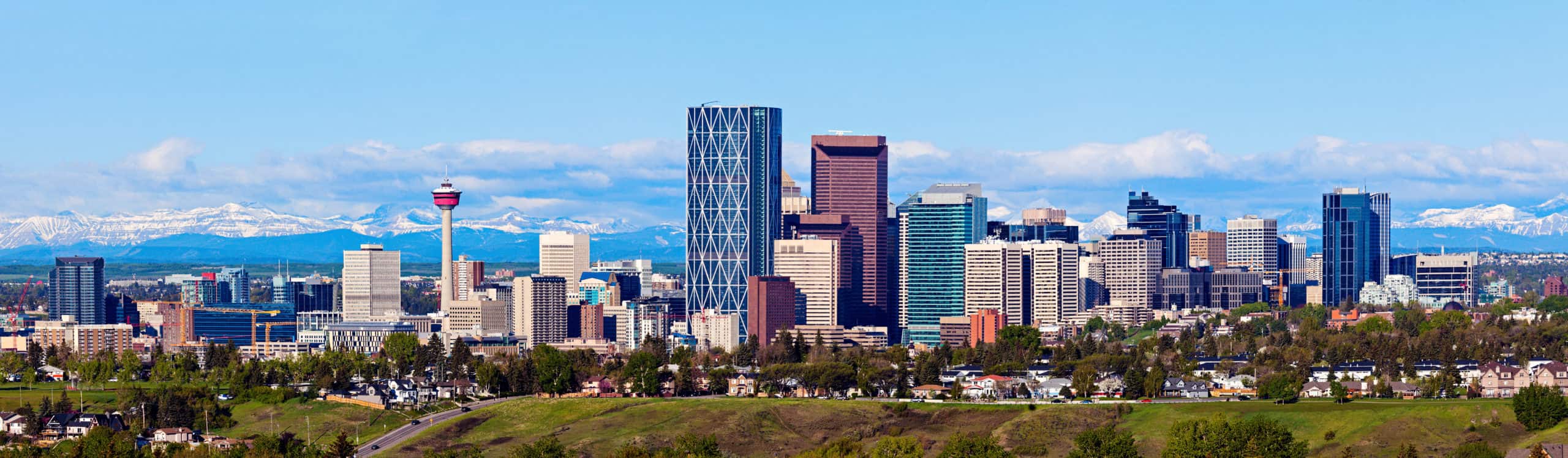 City skyline with modern skyscrapers, red observation tower, green fields in foreground, mountains and blue sky in backdrop.