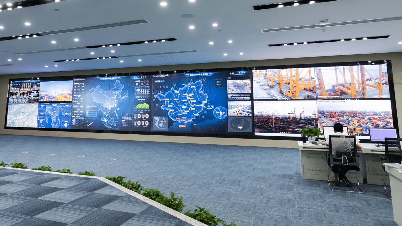 A control room with a digital screen wall; a person monitors data at a desk, carpeted floor, plants along the edge.