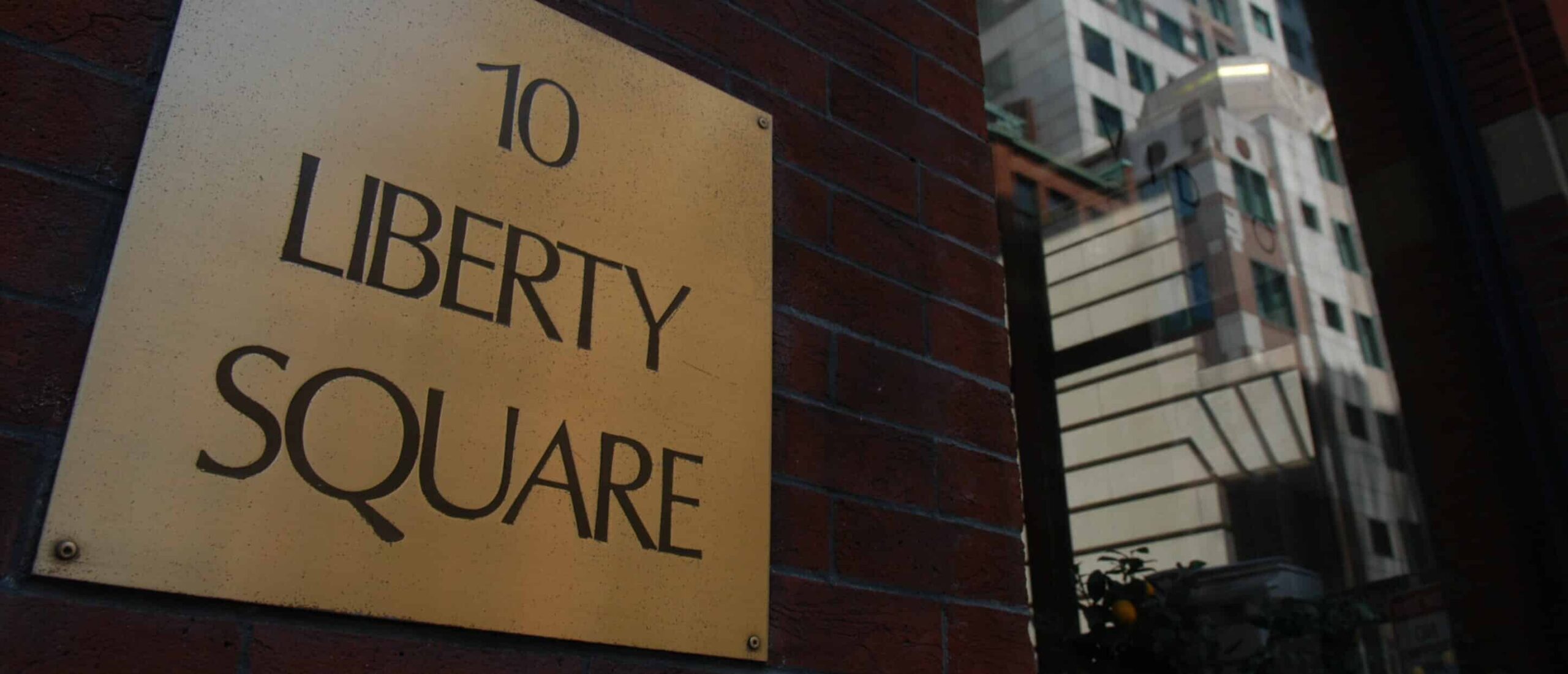 Plaque on brick wall: 10 Liberty Square. Reflections of tall buildings visible in nearby glass.