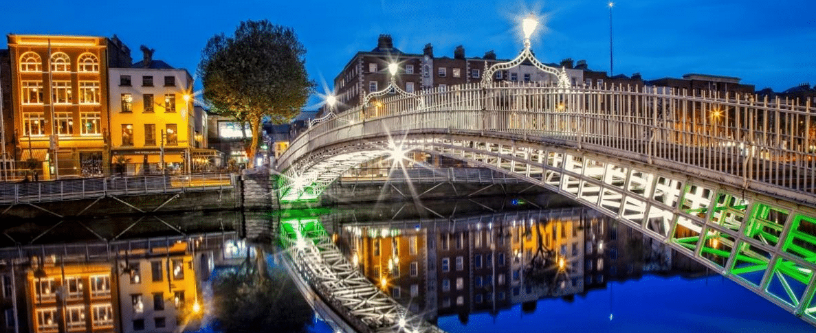 A lit-up bridge with ornate railings spans a calm river, reflecting brightly. Buildings line the bank; a large tree stands nearby.