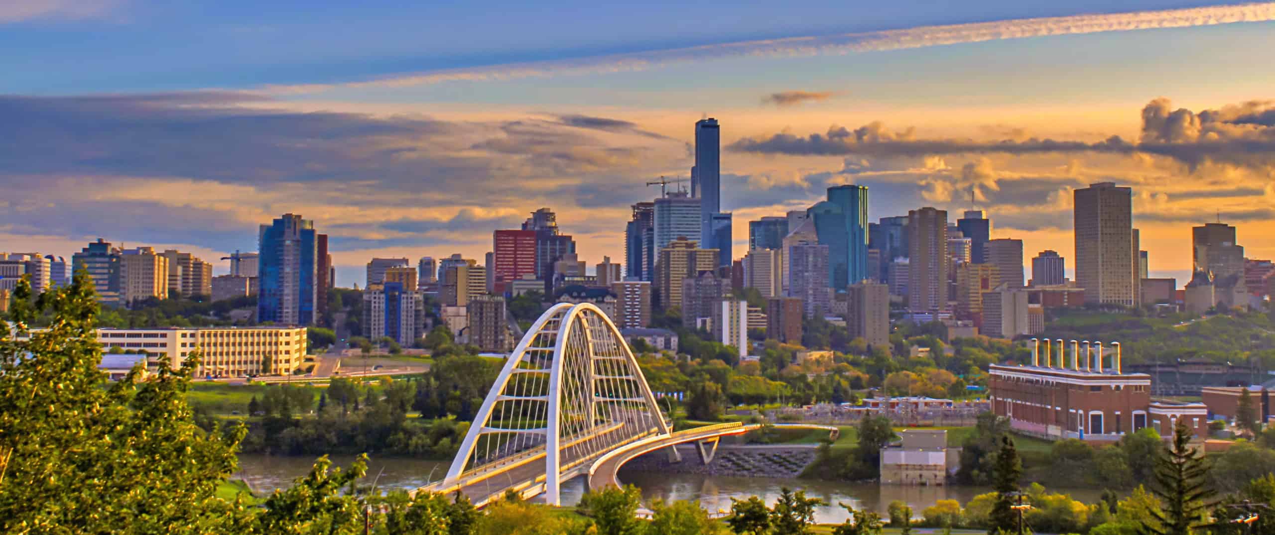 Skyline at sunset with a white arched bridge, skyscrapers, clouds in pastel hues, and lush greenery surrounding the city.