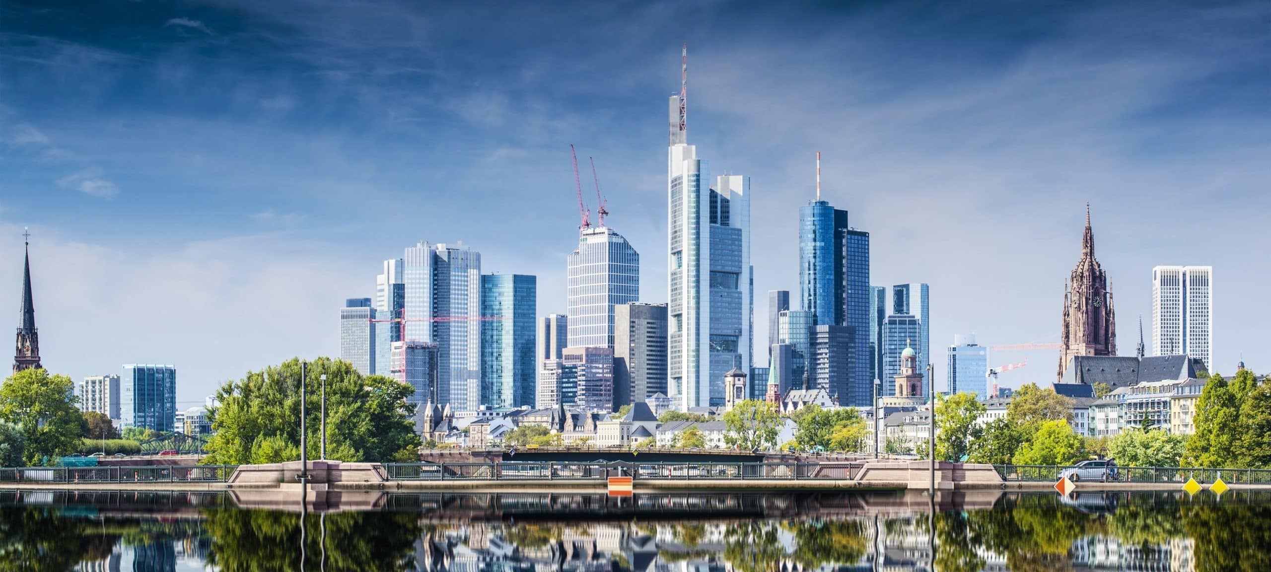 City skyline with skyscrapers and church towers under a blue sky; river reflections and trees framing the view.