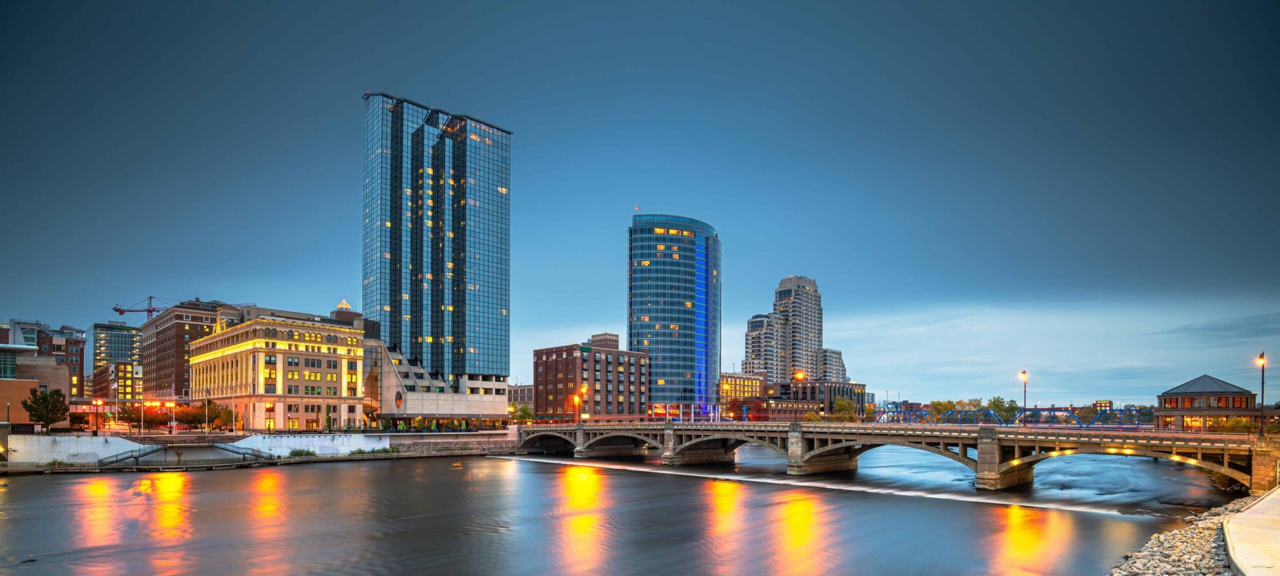 City skyline with modern and historic buildings reflecting in a river at dusk, bridge crossing, illuminated by streetlights under a clear blue sky.