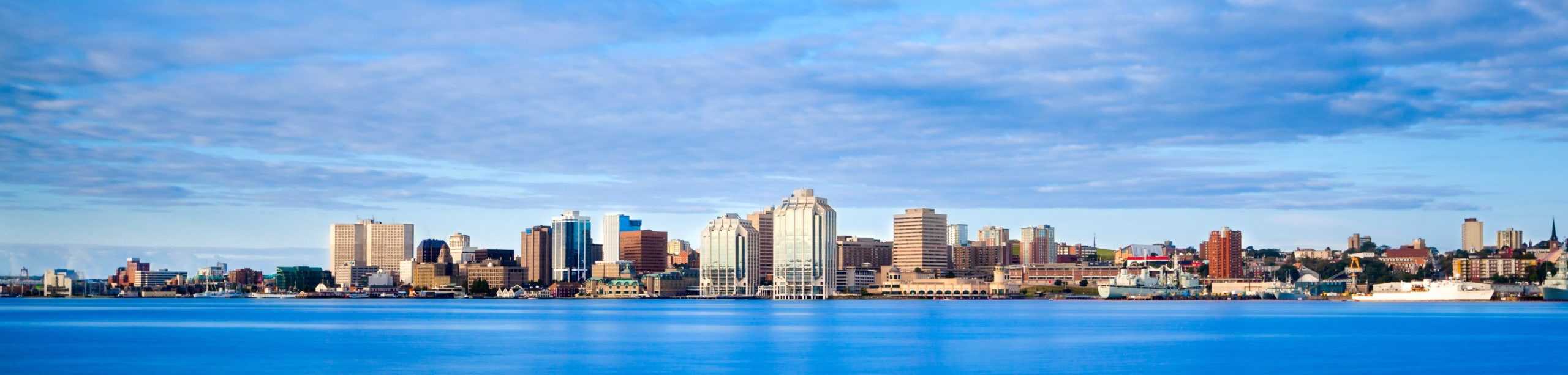 City skyline with tall buildings reflecting in calm coastal waters under a partly cloudy sky.