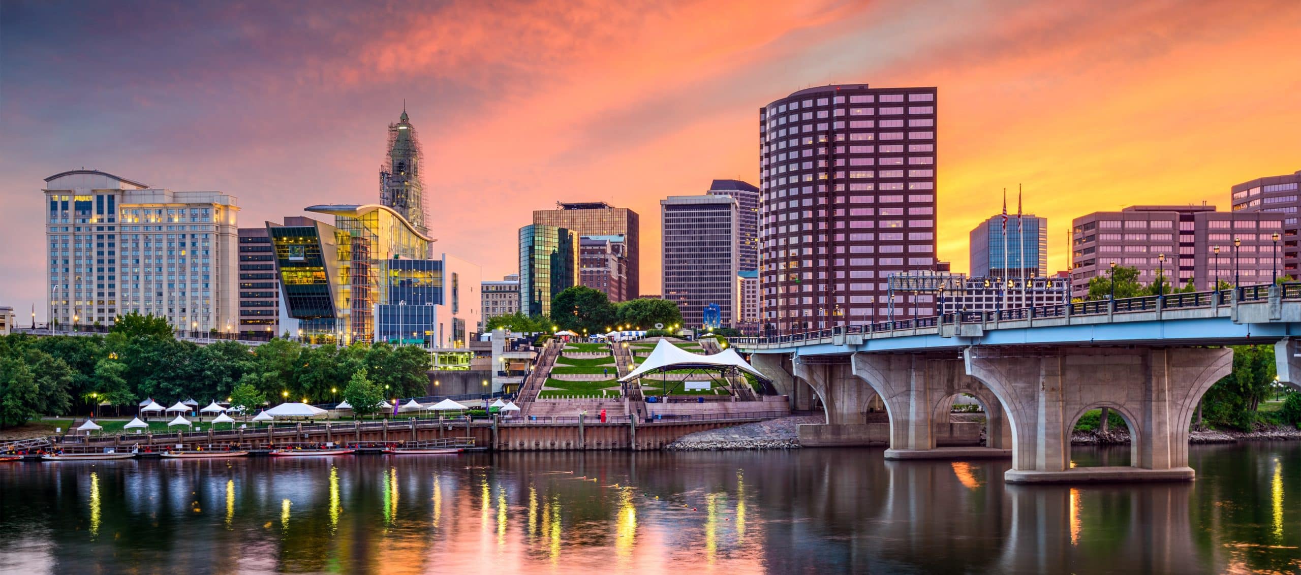 Cityscape at sunset: skyscrapers, orange-pink sky, river with bridge, tents by green riverbank.