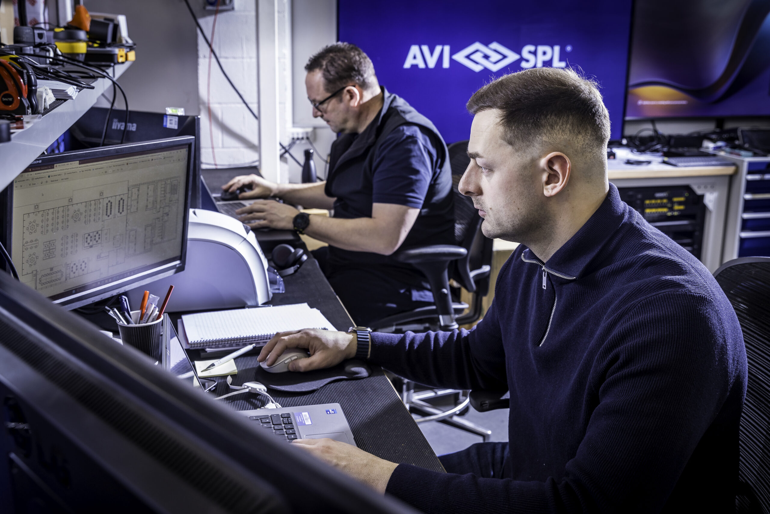 Two men at AVI SPL office, one reviews a blueprint on screen, the other types. Office equipment and supplies are visible.