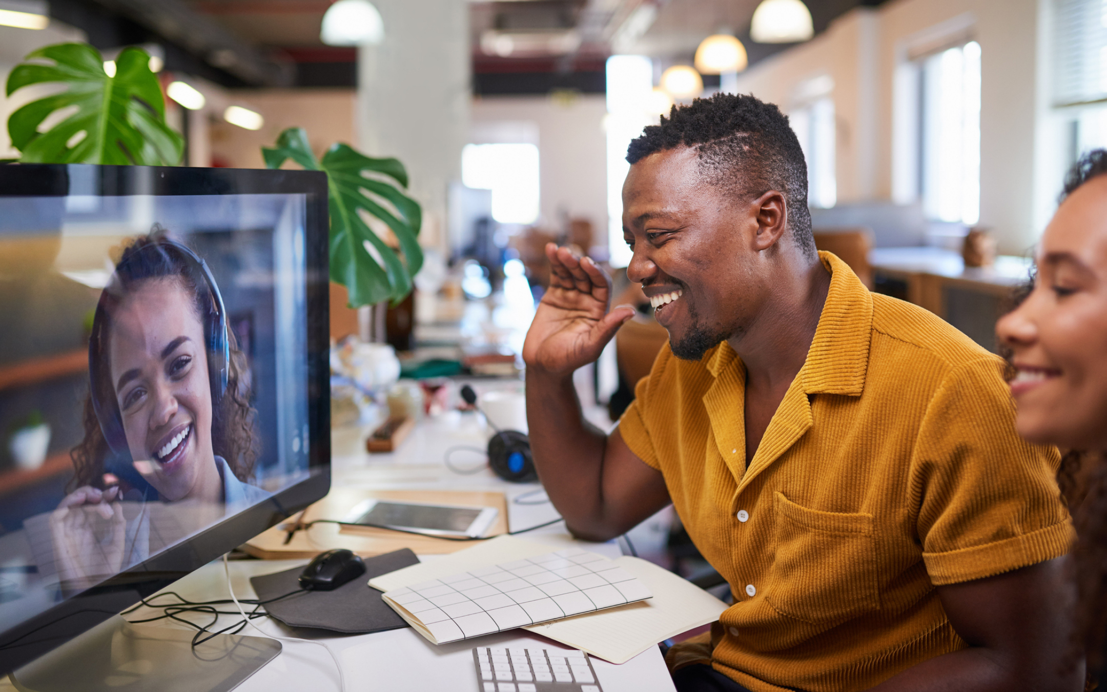 A man in a yellow shirt video calls a woman with headphones; both smile brightly in a modern office, joined by another smiling woman.