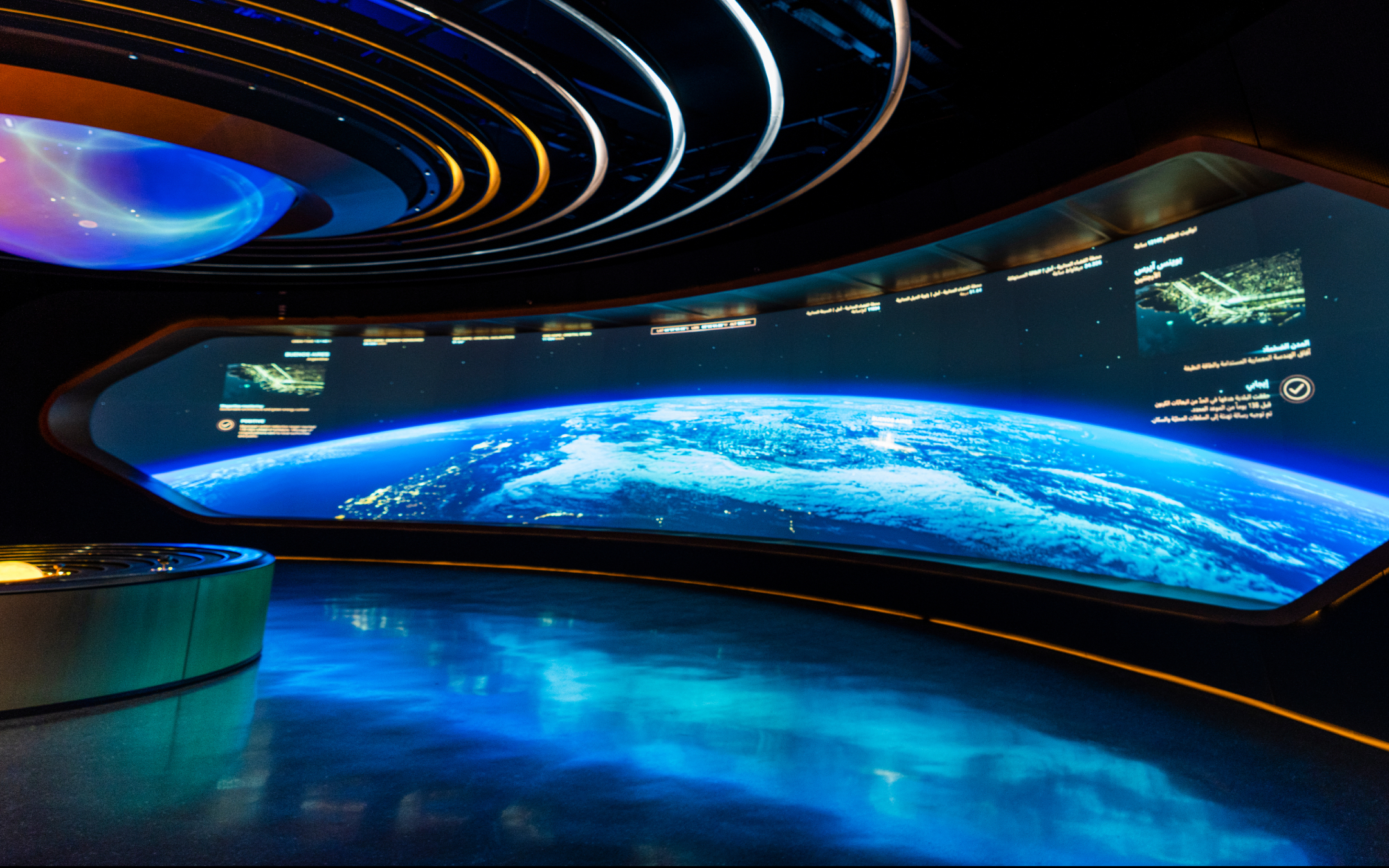 A futuristic room has a curved screen showing Earth from space. Blue-glowing floor, circular neon lights complete the sleek look.