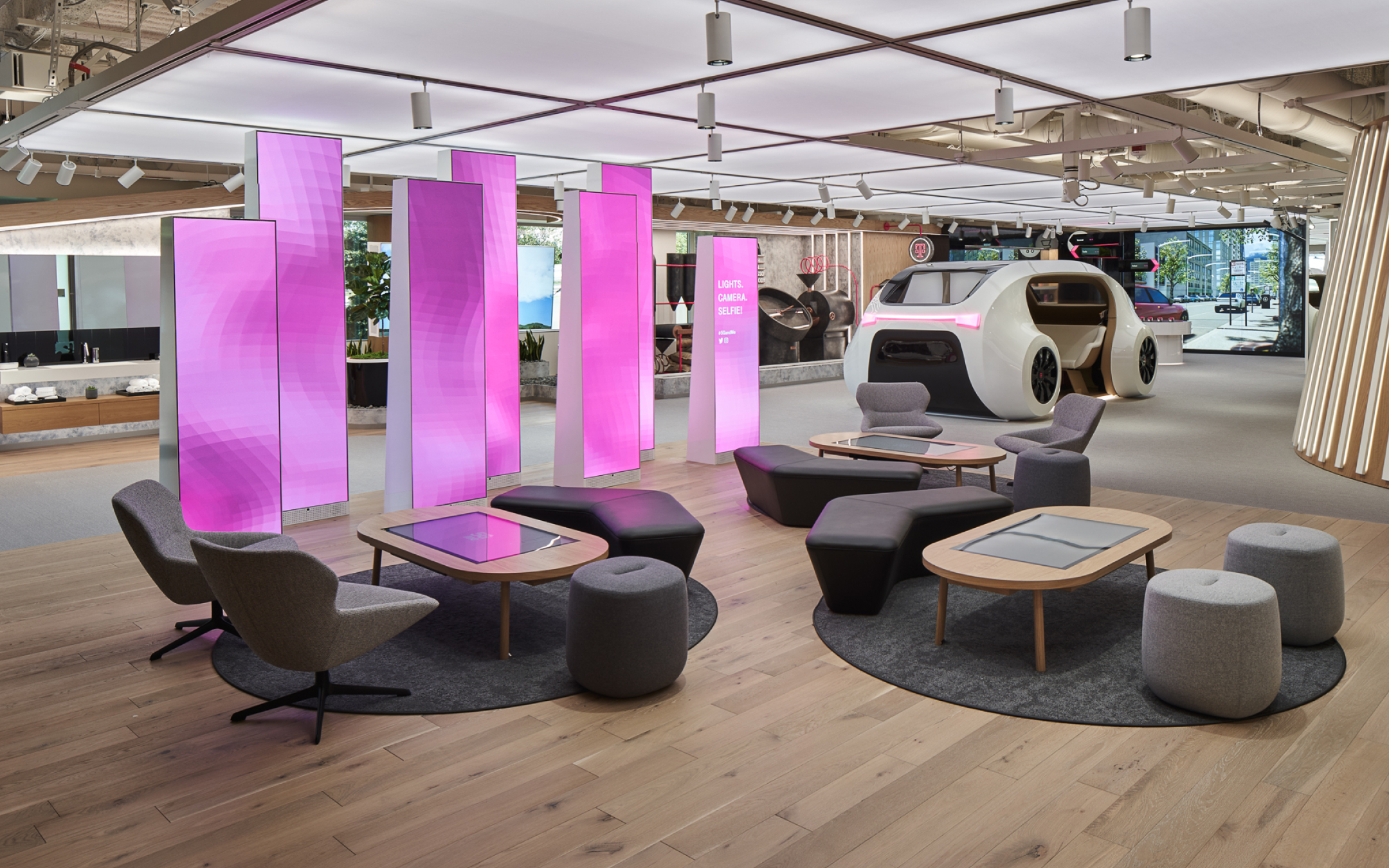 A modern showroom with pink screens, contemporary furniture on circular rugs, a futuristic vehicle, and bright inviting lighting.