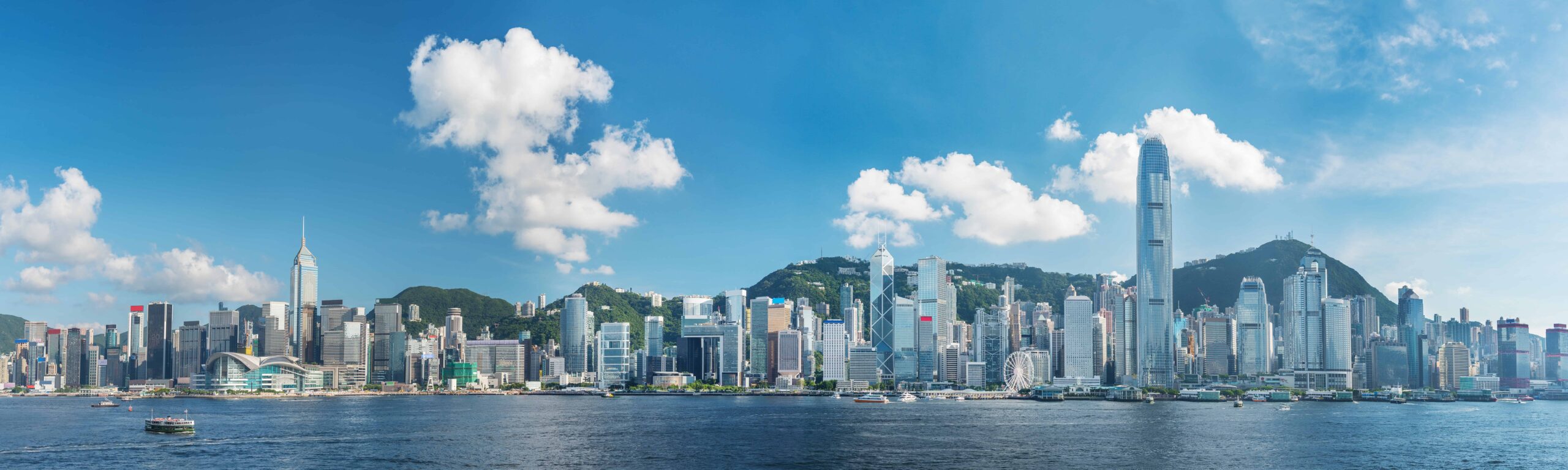 Panoramic Hong Kong skyline: skyscrapers, blue sky with clouds, Victoria Harbour and boats in foreground, hills in background.