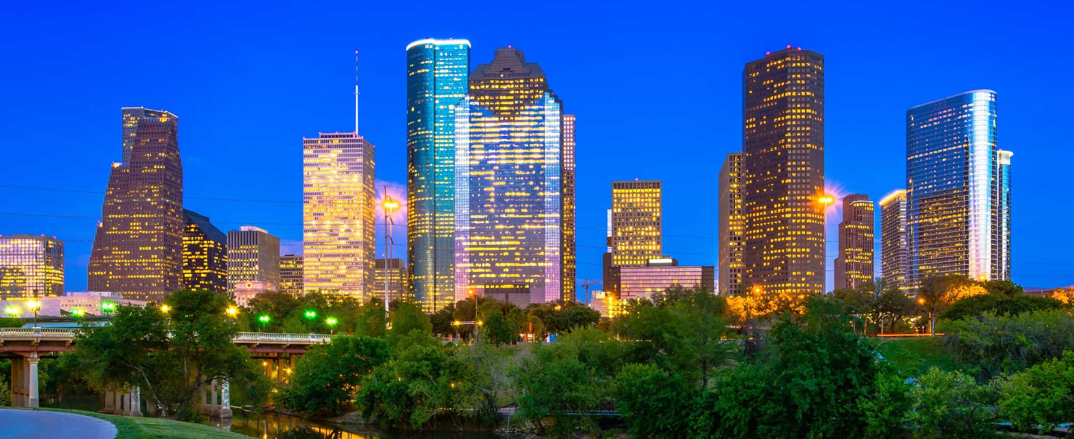 Nighttime city skyline with lit skyscrapers, deep blue sky, green trees, and a reflective waterway in the foreground.