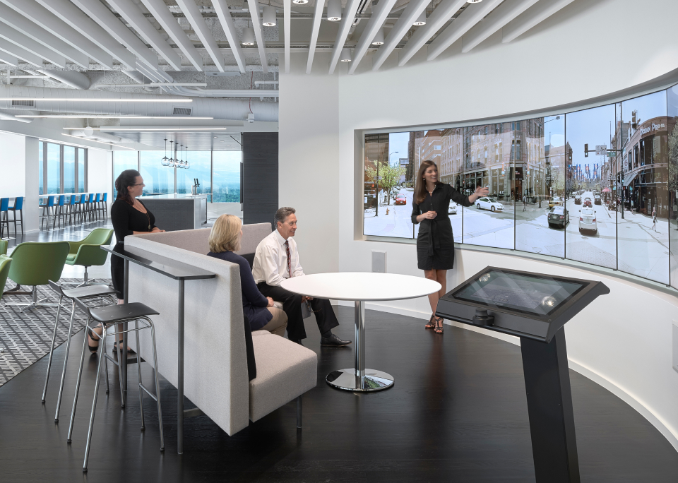 A woman presents in a modern office with a curved screen; three colleagues listen attentively amid sleek, contemporary decor.