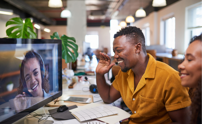 Man in yellow shirt waves at woman on video call; another person smiles beside him in a well-lit office with plants and notebooks.