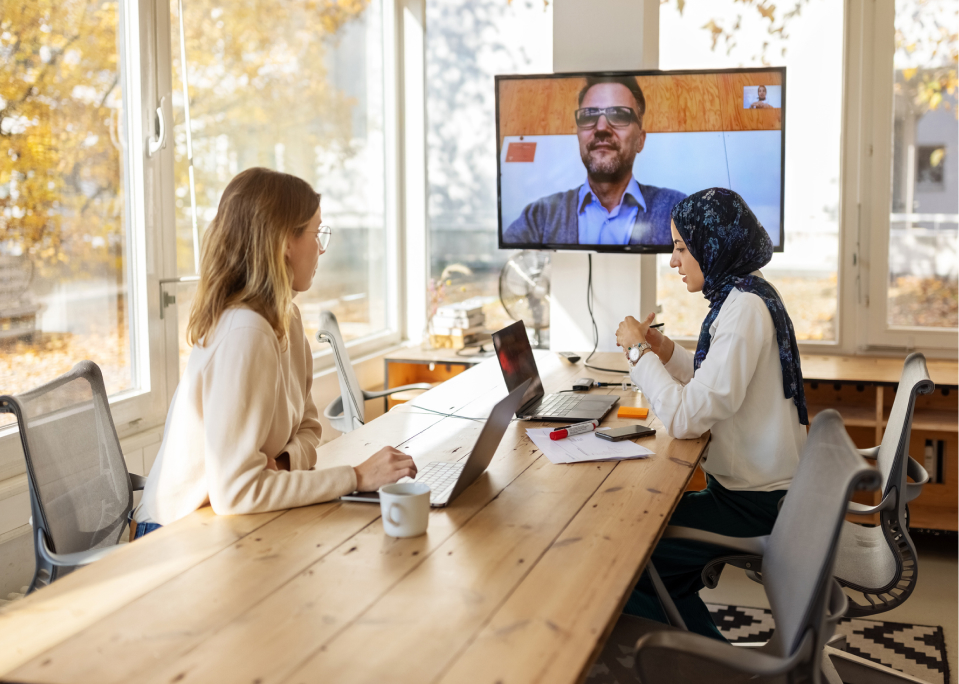 Two women at a table with laptops video conference with a man on screen; large windows reveal trees. Papers and mug on table.