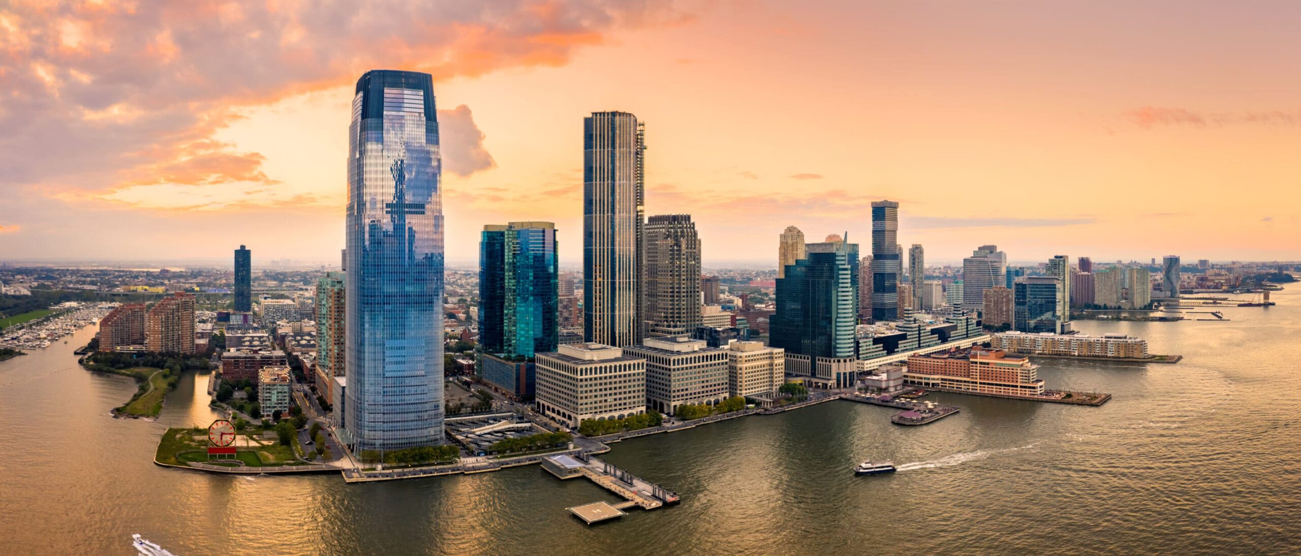 Aerial view: Modern cityscape at sunset, glass skyscrapers reflecting sky, calm river with boats, vibrant orange horizon.