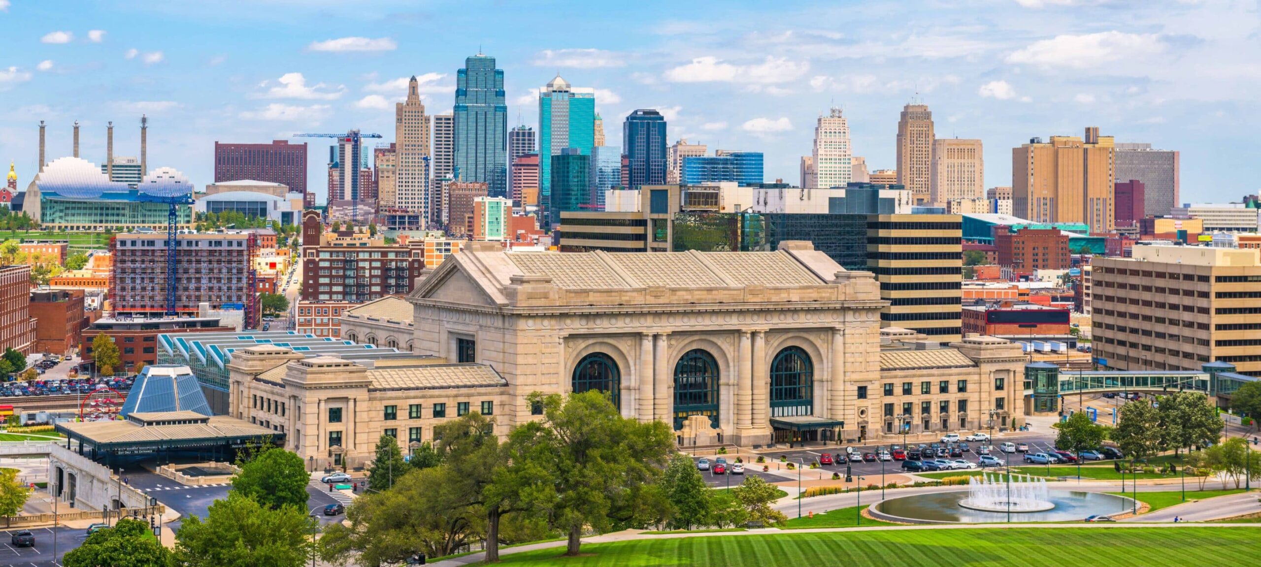 Cityscape aerial view: historic building with arched windows, modern skyscrapers, park with fountain, lush greenery, partly cloudy sky.