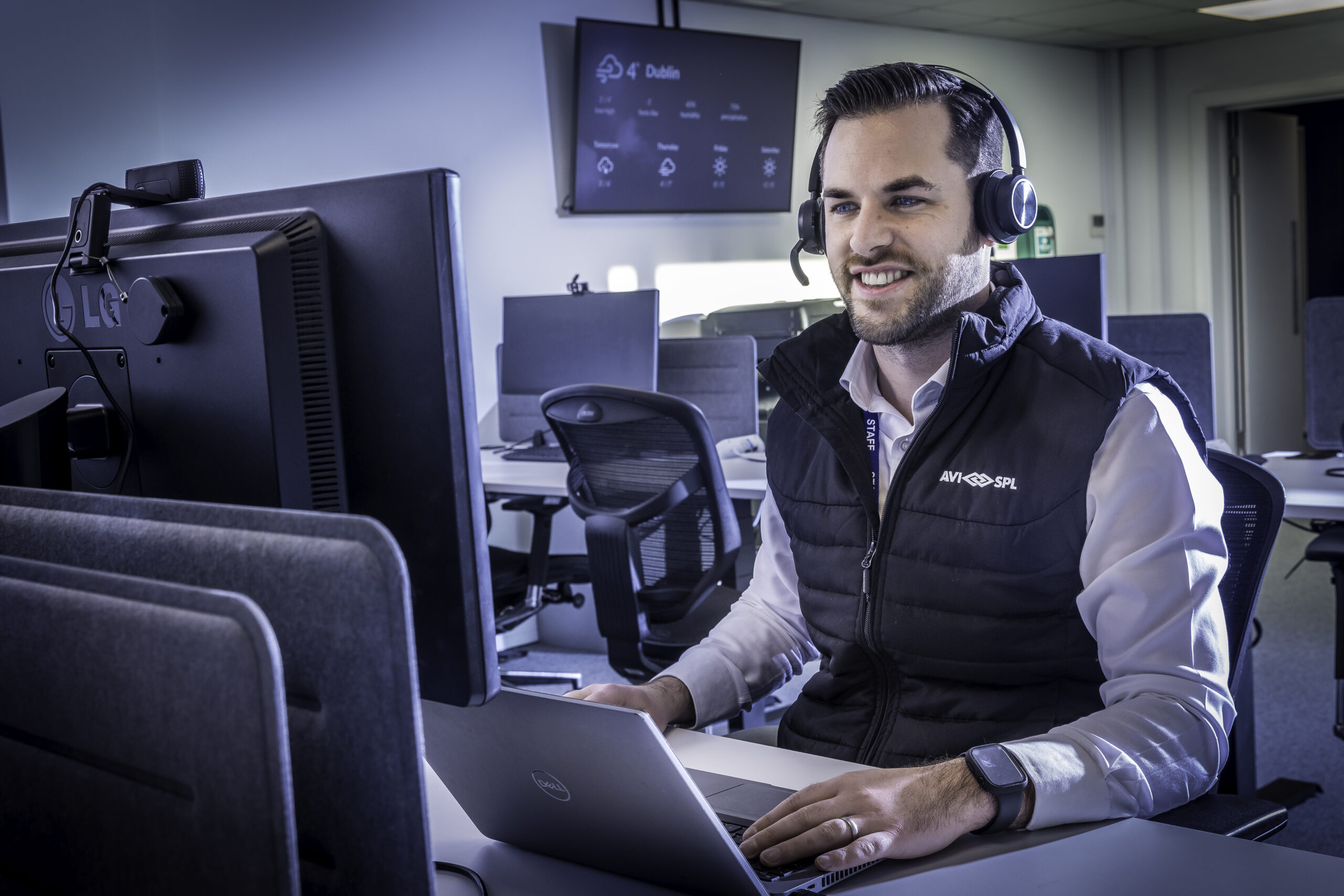 A person in a headset and black vest smiles while working at a computer, with multiple monitors and a screen showing Dublin.