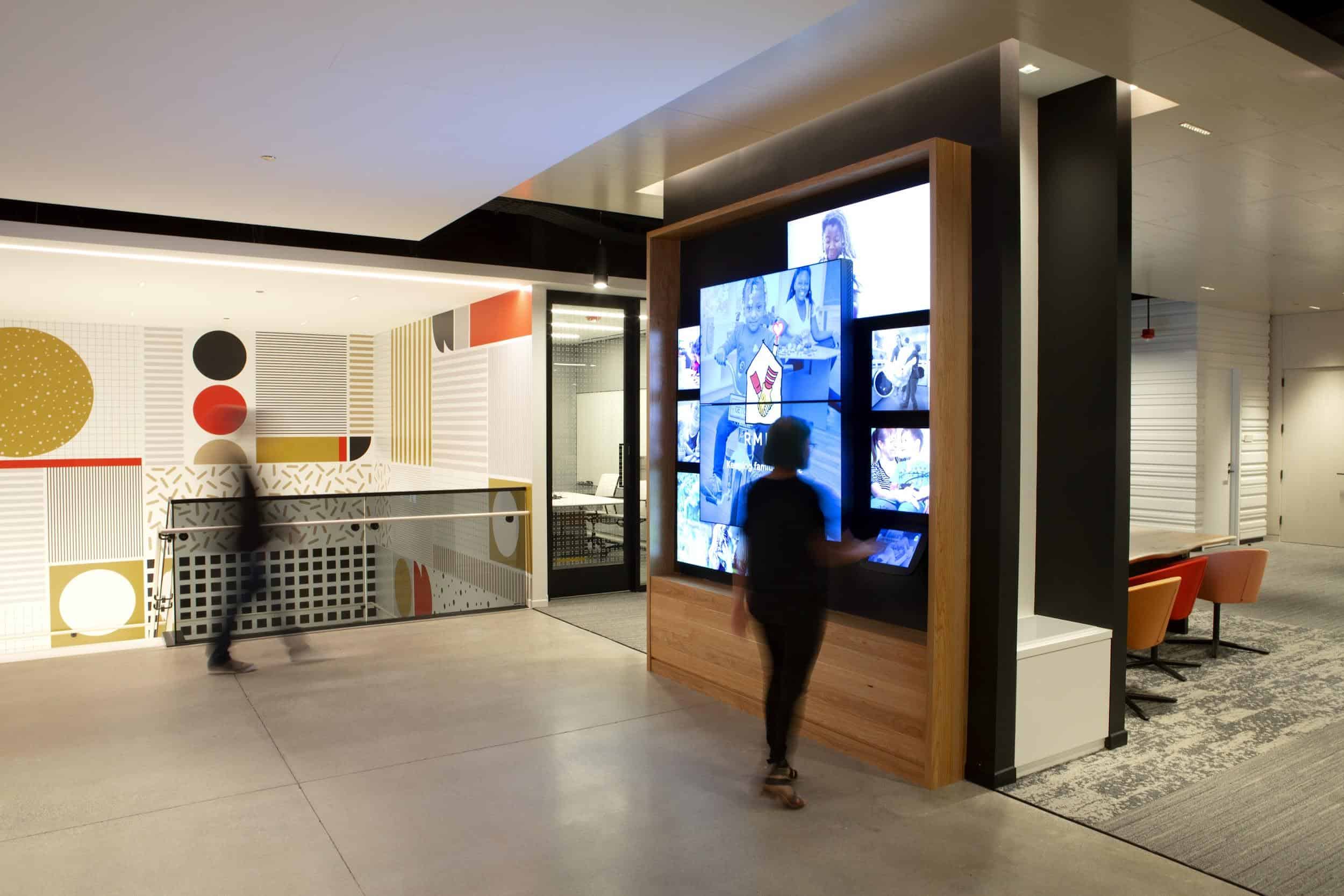 Modern office with digital display wall, geometric decor in red, black, gold; blurred figures; open space with seating and work areas.