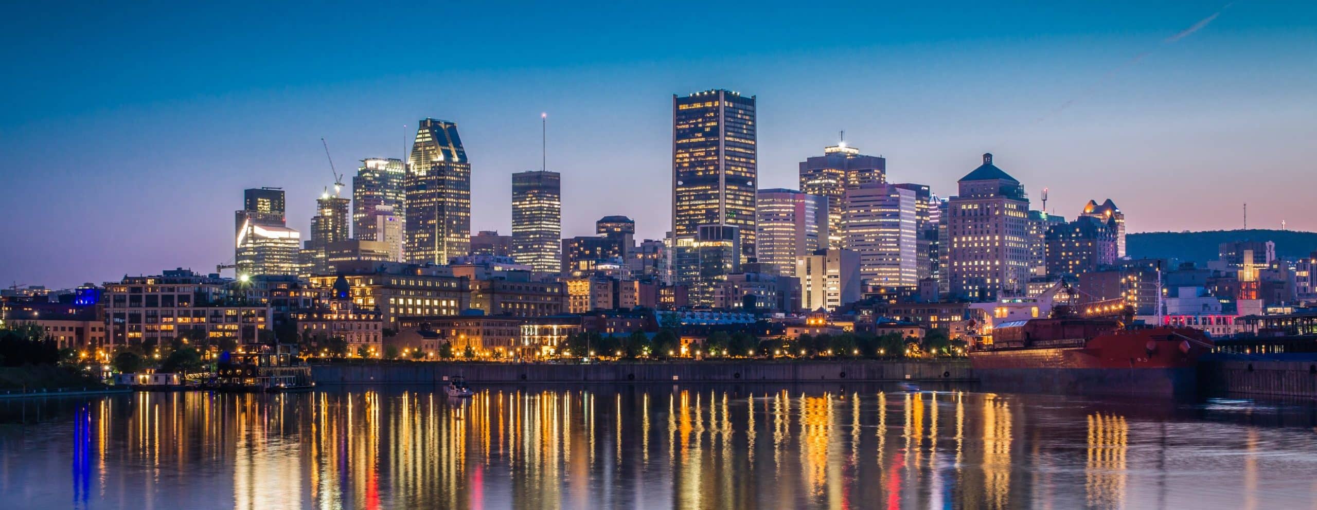 City skyline at dusk with lit skyscrapers mirrored in calm water; sky fades deep to light blue, lights reflecting vibrantly.