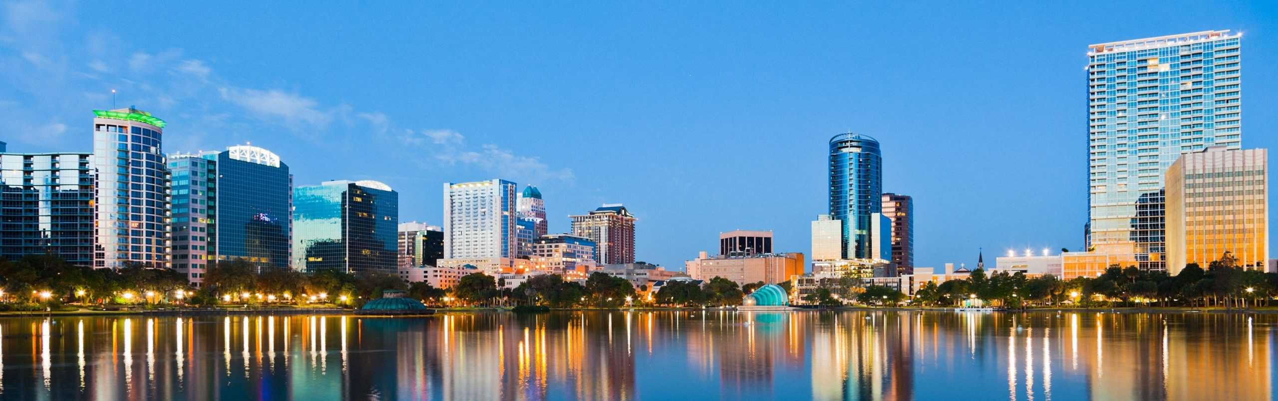 Modern city skyline at dusk, tall lit buildings mirrored in calm water, trees along the edge; a peaceful, vibrant scene.