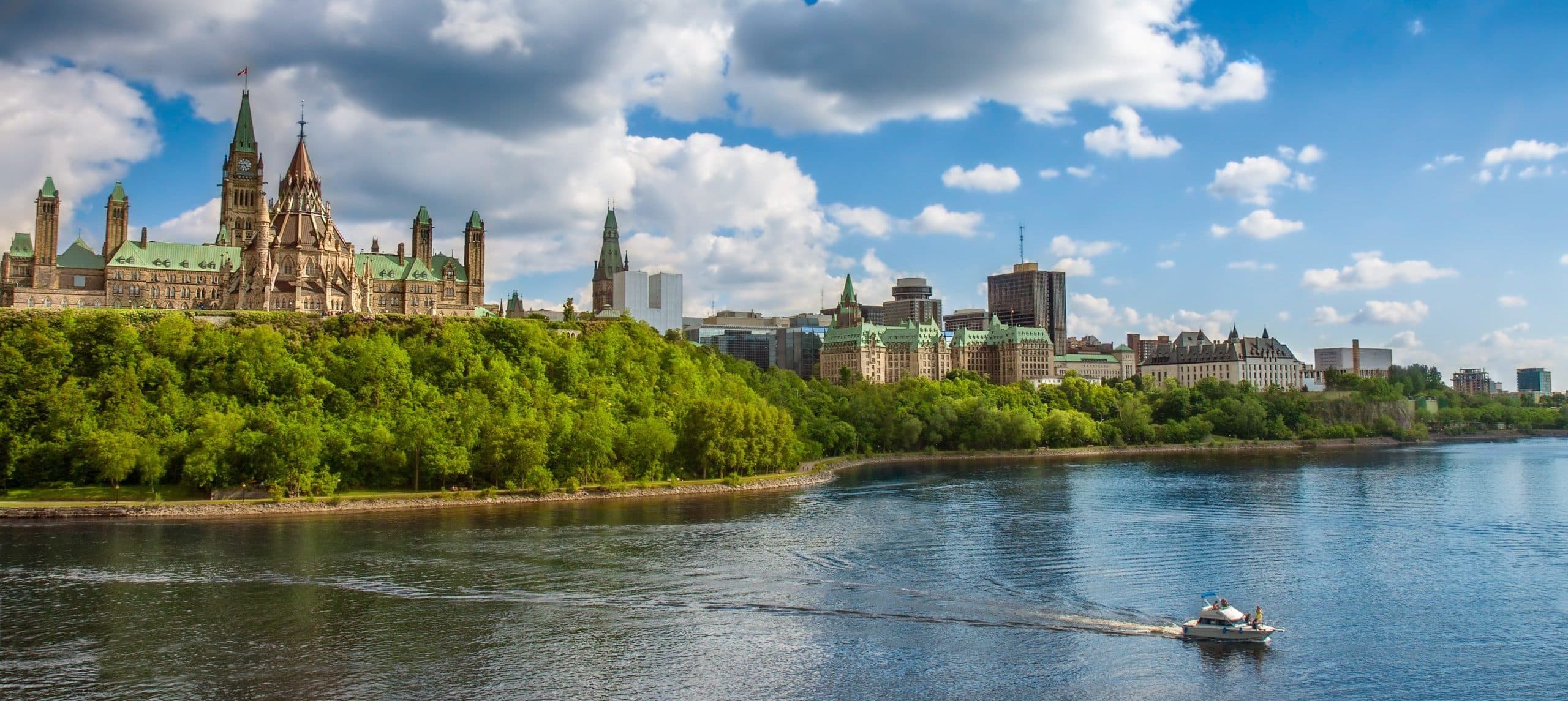 Ottawas Parliament Hill with green roofs and spires, lush trees. Boat on Ottawa River under partly cloudy blue sky.