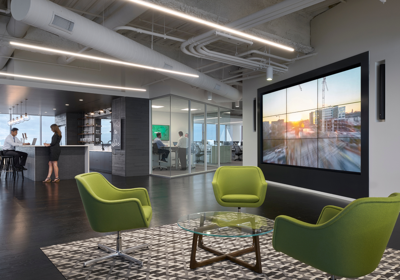 Modern office: green chairs, glass table, patterned rug, cityscape screen. People converse at bar; others work in glass rooms. Exposed ceiling.