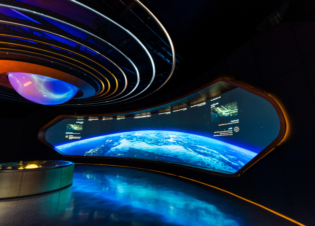 Futuristic room: large curved screen showing Earth, blue/gold lights, concentric ceiling circles, round display table in front.