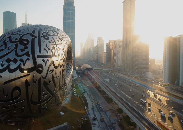 Aerial view of futuristic cityscape with oval building featuring Arabic calligraphy, tall skyscrapers, and busy highway.