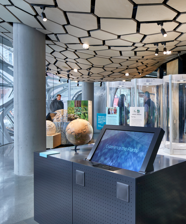 Futuristic exhibit: touchscreen Experience the Plants, globe, cylinders; geometric ceiling, natural light, 2 observers.